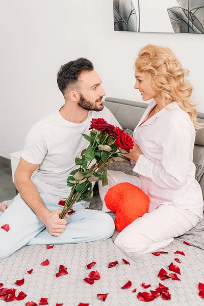 Blissful couple sitting on bed with roses and toy heart — Stock Photo