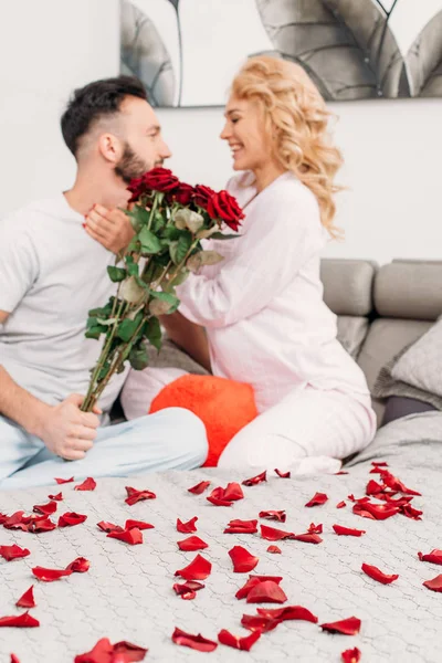 Smiling couple with red roses sitting on bed and looking at each other — Stock Photo