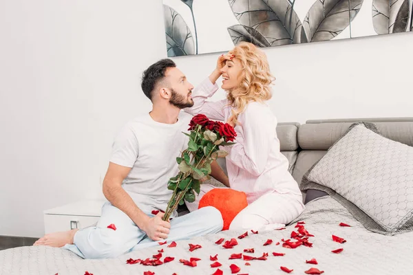 Smiling romantic couple sitting on bed with red roses — Stock Photo