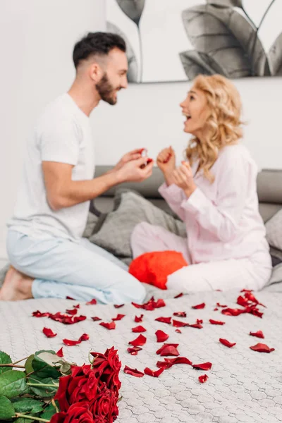 Мужчина с бородой сидит на кровати с лепестками роз и делает предложение девушке — стоковое фото