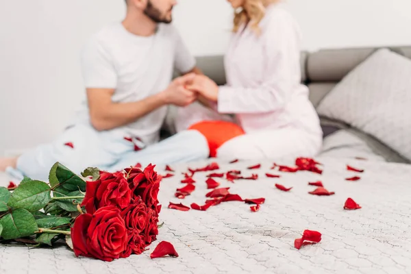 Vista parcial de la pareja sentada en la cama con pétalos de rosa y cogidas de la mano - foto de stock