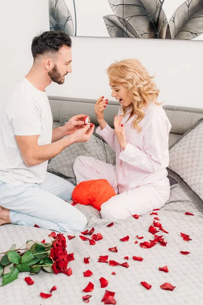 Man sitting on bed with rose petals and proposing to curly girl — Stock Photo