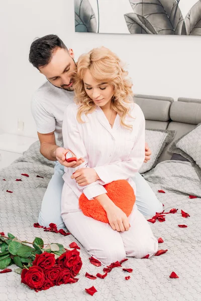 Handsome man sitting on bed with roses and proposing to attractive woman — Stock Photo