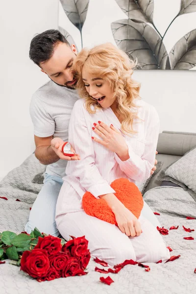 Hombre sentado en la cama con rosas rojas y proponiéndole matrimonio a su novia sorprendida - foto de stock