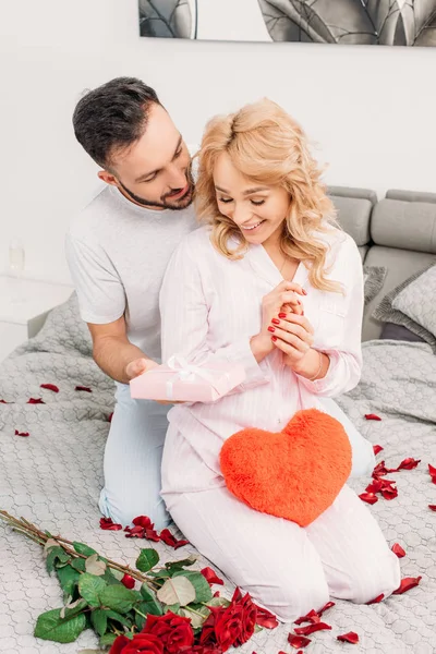 Smiling man sitting on bed with roses and presenting gift to girlfriend — Stock Photo