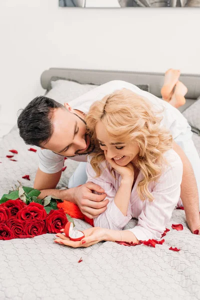 Blissful couple lying on bed with ring and red roses — Stock Photo