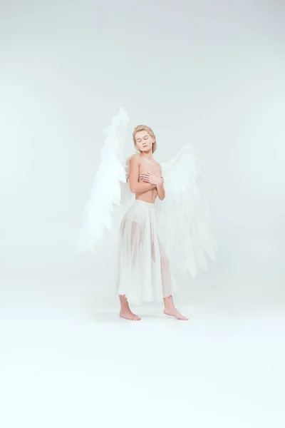 Hermosa mujer con alas de ángel cubriendo los pechos y posando con los ojos cerrados aislados en blanco - foto de stock
