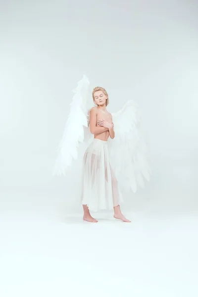 Bela mulher com asas de anjo cobrindo seios e posando com os olhos fechados isolado no branco — Fotografia de Stock