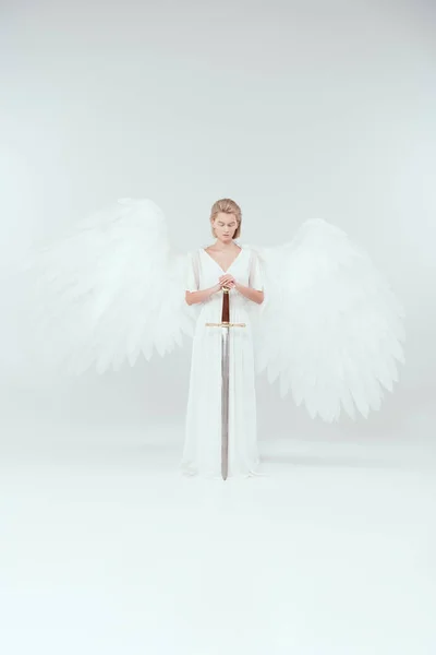 Hermosa mujer con alas de ángel sosteniendo la espada y posando sobre fondo blanco - foto de stock