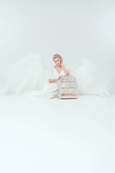 Hermosa mujer tierna en traje de ángel con alas sentado y posando con jaula de pájaro aislado en blanco - foto de stock
