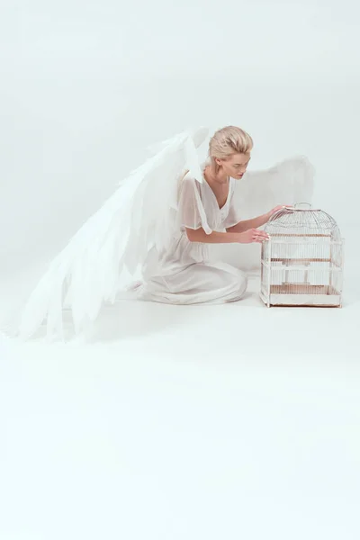 Hermosa mujer en traje de ángel con alas posando con jaula de pájaro aislado en blanco - foto de stock