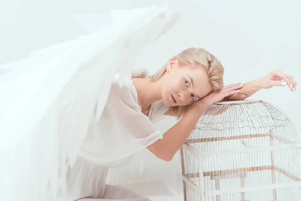 Foco seletivo da mulher bonita em traje de anjo com asas olhando para a câmera e inclinando-se na gaiola de pássaro isolado no branco — Fotografia de Stock