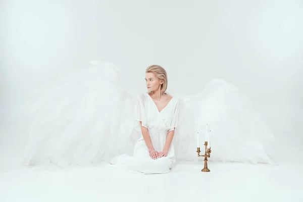 Красивая женщина в ангельском костюме с крыльями, сидящая возле канделябра со свечами и смотрящая в сторону изолированной на белом — стоковое фото
