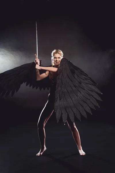 Belle femme en colère en costume de guerrier avec des ailes d'ange tenant épée et posant sur fond noir — Photo de stock