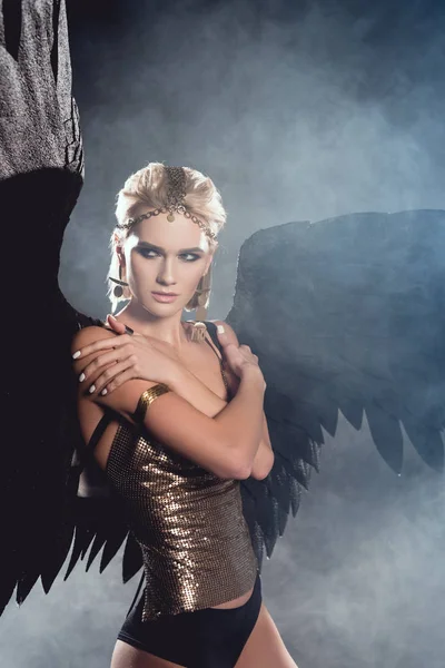 Красива молода сексуальна жінка з чорними крилами ангела і золотими аксесуарами позує на темному димчастому фоні — стокове фото