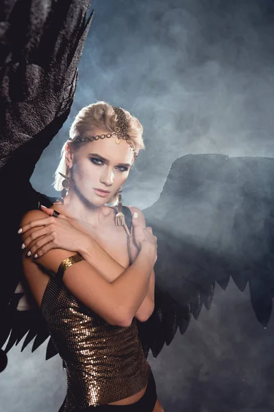 Beautiful young sexy woman with black angel wings and golden accessories posing on dark smoky background — Stock Photo