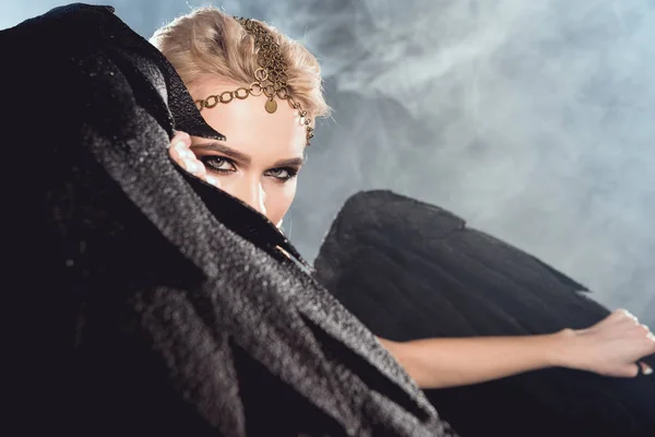 Belle femme sexy dans des accessoires dorés tenant des ailes d'ange noir et posant sur fond sombre — Photo de stock