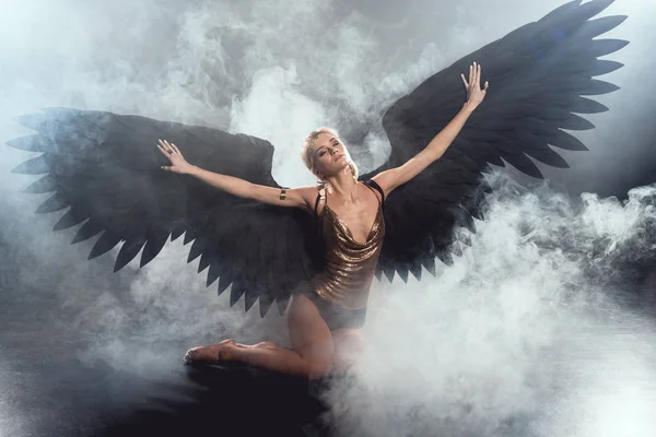 Красивая сексуальная женщина с черными крыльями ангела и протянутыми руками сидя и позируя на темном дымном фоне — стоковое фото