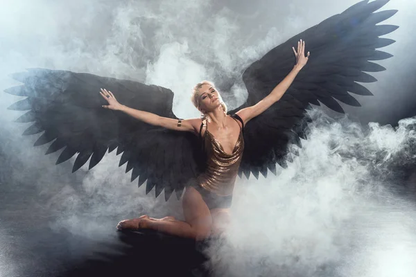 Beautiful sexy woman with black angel wings and outstretched hands sitting and posing on dark smoky background — Stock Photo