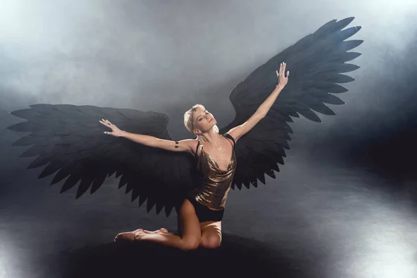 Красивая сексуальная женщина с черными крыльями ангела и протянутыми руками сидя и позируя на темном фоне — стоковое фото