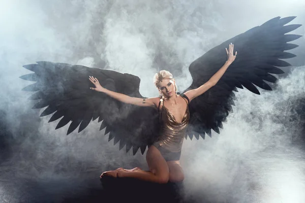 Belle femme sexy avec des ailes d'ange noir et les mains tendues assis et posant sur fond sombre fumé — Photo de stock