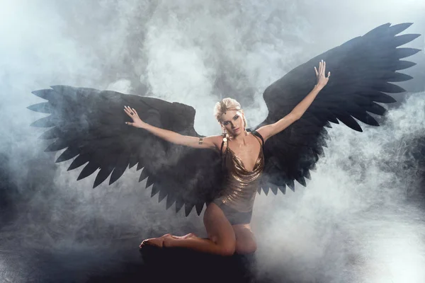 Beautiful sexy woman with black angel wings and outstretched hands sitting and posing on dark smoky background — Stock Photo