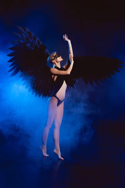 Красивая сексуальная женщина с кружевной маской и черными крыльями ангела стоя на цыпочках и позируя на темно-синем фоне — стоковое фото