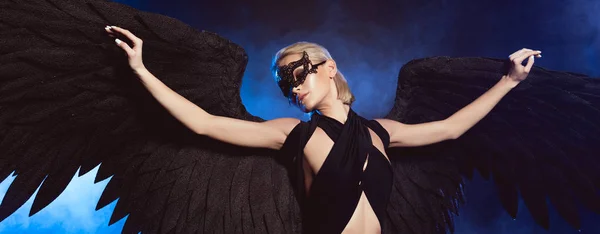 Красива сексуальна жінка з мереживною маскою і чорними ангельськими крилами позує на темно-синьому фоні — стокове фото