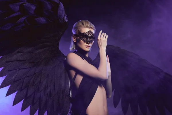 Beautiful sexy woman in lace mask and black angel wings gesturing with hand and posing on violet background — Stock Photo