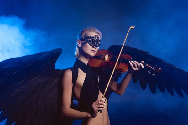 Schöne sexy Frau mit Spitzenmaske und schwarzen Engelsflügeln spielt Geige auf dunkelblauem rauchigen Hintergrund — Stockfoto
