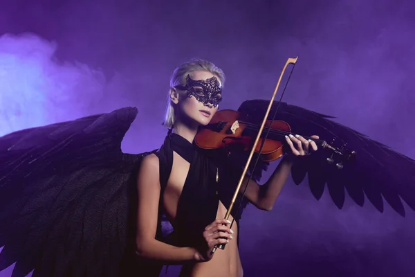 Beautiful woman in lace mask and black angel wings playing fiddle on violet background — Stock Photo