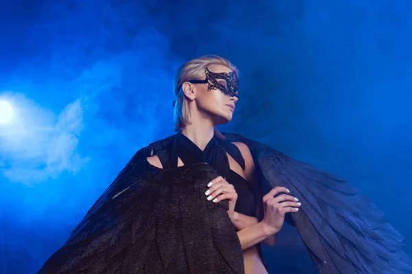 Hermosa mujer sexy en máscara de encaje sosteniendo alas de ángel negro y posando sobre fondo azul oscuro - foto de stock