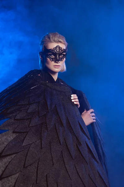 Beautiful mysterious woman with lace mask and black angel wings looking at camera on dark blue background — Stock Photo