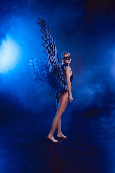 Hermosa mujer misteriosa sexy en máscara de encaje y alas de ángel negro posando sobre fondo ahumado azul oscuro — Stock Photo