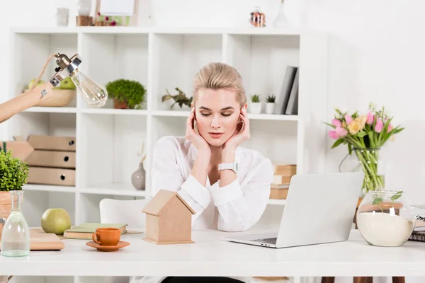 Привлекательная женщина смотрит на небольшой картонный домик, сидя рядом с ноутбуком в офисе, концепция сохранения окружающей среды — стоковое фото