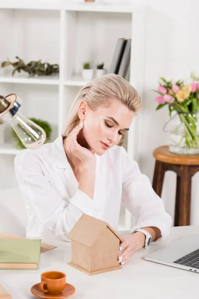 Attraktive Frau blickt auf kleines Papphaus, während sie neben Tasse mit Getränk sitzt, Umweltschutzkonzept — Stockfoto