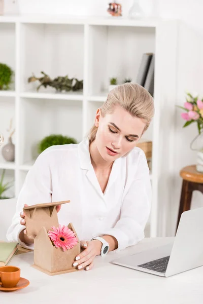 Mulher atraente olhando para a flor rosa em pequena casa de papelão enquanto sentado perto do laptop no escritório, conceito de poupança ambiental — Fotografia de Stock