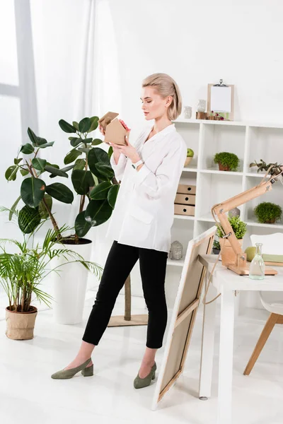 Donna d'affari che tiene la piccola casa di cartone con fiore vicino a piante verdi in ufficio, concetto di risparmio ambientale — Foto stock