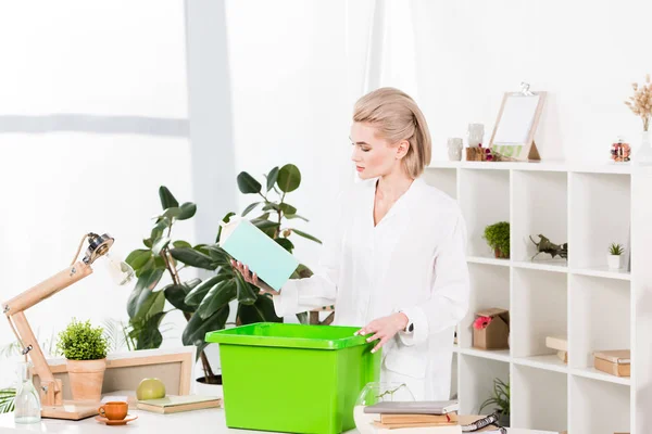 Attractive woman holding milk package near recycling box while standing in office, environmental saving concept — Stock Photo