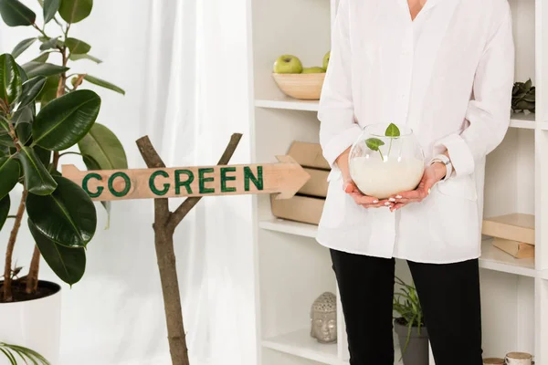 Cropped view of woman holding glass fish bowl with sand and green leaves near go green sign in office, environmental saving concept — Stock Photo