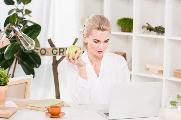 Attractive woman looking at laptop and holding green apple in office — Stock Photo