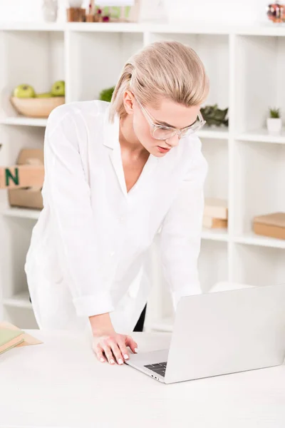 Atractiva mujer en gafas mirando a la computadora portátil mientras está de pie en la oficina - foto de stock