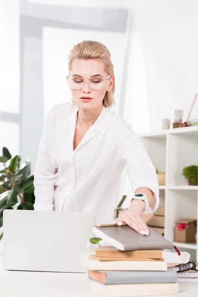 Atractiva mujer de negocios en gafas tomando libro de pie cerca de la computadora portátil en la oficina - foto de stock