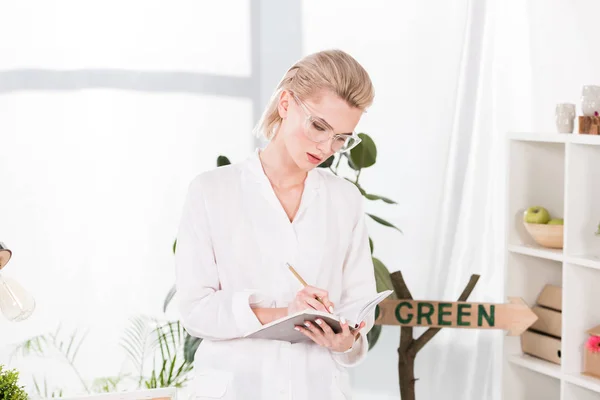 Pensativa mujer de negocios en gafas de escribir en cuaderno mientras está de pie en la oficina, concepto de ahorro ambiental - foto de stock