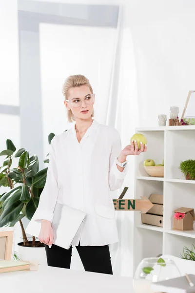 Attraente donna in piedi con computer portatile e tenendo mela verde in ufficio — Foto stock