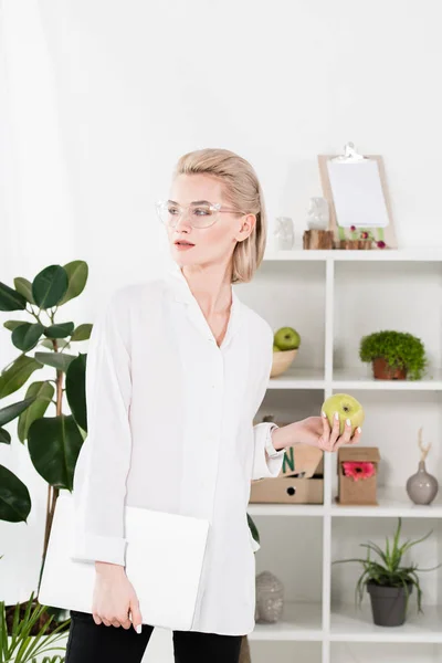 Belle femme dans des lunettes debout avec ordinateur portable et pomme verte au bureau — Photo de stock