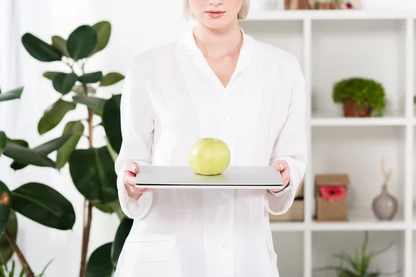 Cropped view of businesswoman holding laptop with green apple in office — Stock Photo