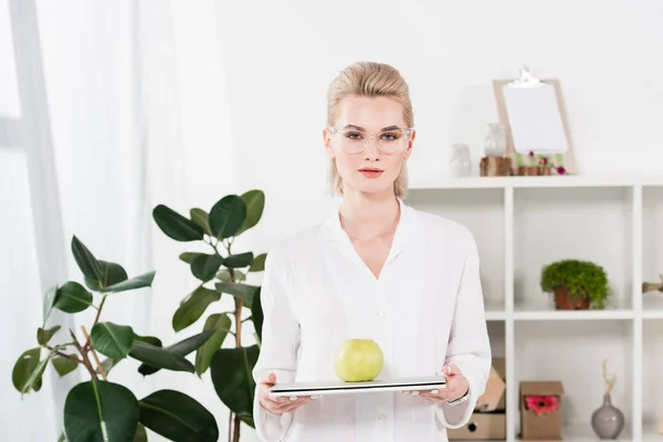 Donna d'affari attraente in occhiali che tengono il computer portatile con mela verde in ufficio — Foto stock