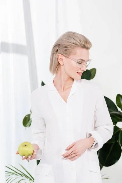 Cheerful businesswoman in glasses holding green apple in office — Stock Photo