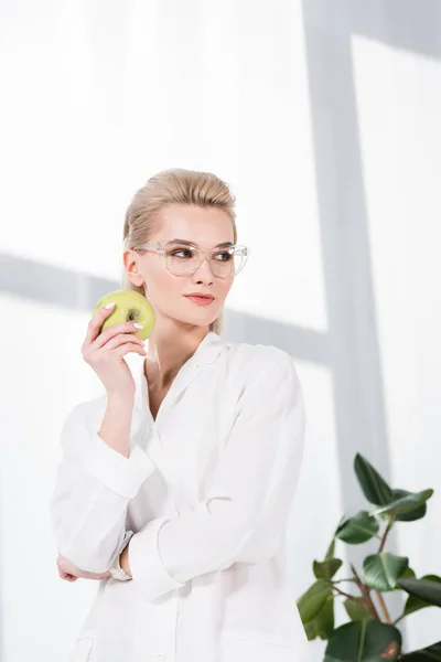 Smiling businesswoman in glasses holding green apple in office — Stock Photo